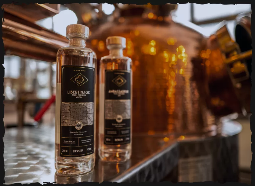 Flaschen von Babylon Berlin Libertinage Vodka und Ambassadeur Gin vor einer Kupfer-Destillerie